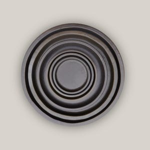 May include: A dark grey, circular object with concentric rings, creating a layered effect. The centre is a small, flat circle. The overall design is simple and modern, with a slightly textured appearance. The object appears to be made of metal.