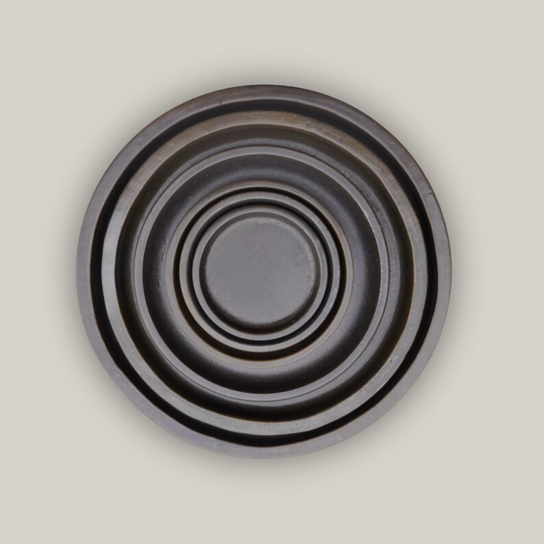 May include: A dark grey, circular object with concentric rings, creating a layered effect. The centre is a small, flat circle. The overall design is simple and modern, with a slightly textured appearance. The object appears to be made of metal.