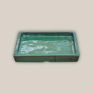 May include: A rectangular, glazed ceramic tray in a vibrant teal green. The tray has a slightly raised rim and a smooth, reflective surface. The edges are a darker shade of green, adding depth to the design. This tray is suitable for home decor.