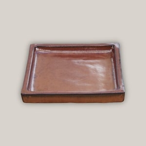 May include: A square, terracotta-colored ceramic tray with a slightly raised rim. The tray has a smooth, glossy finish and is perfect for serving snacks or displaying small items.