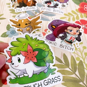 May include: A set of five stickers featuring cartoon characters from the Pokemon franchise. The stickers are printed on white vinyl with a glossy finish. The stickers feature the text "DO CRIME", "BASIC BITCH", "TOXIC BITCH", and "GO TOUCH GRASS".