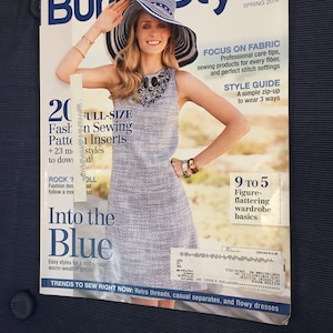 Puede incluir: Una portada de revista para BurdaStyle, con una mujer que lleva un vestido azul claro y un sombrero a rayas blanco y negro. El texto de la portada dice "BurdaStyle" y "Primavera 2014". La portada también presenta el texto "Focus on Fabric", "Style Guide", "9 to 5", y "Into the Blue".