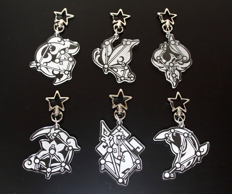 May include: Six black and white keychains with different designs. Each keychain features a star-shaped charm and a unique design with letters and lines. The keychains are on a black background.