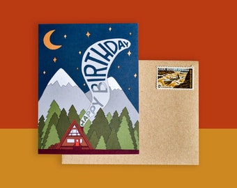 May include: A birthday card with a mountain and forest scene under a starry night sky. The card features the words "Happy Birthday" in a stylized font. An envelope with a postage stamp is included.