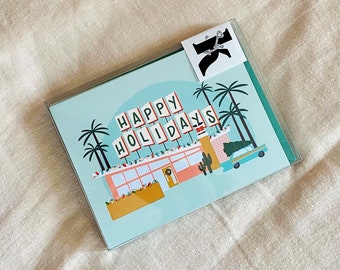 May include: A turquoise blue greeting card with a retro illustration of a diner with palm trees and a car. The diner has a pink and orange exterior and the sign says "Happy Holidays".
