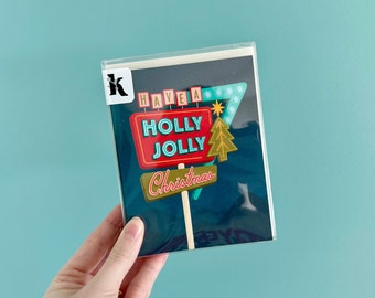 May include: A Christmas card with a retro-style sign that reads "Have a Holly Jolly Christmas". The sign is red and white with blue neon lights and a green Christmas tree.