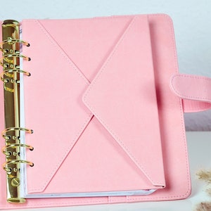 Könnte beinhalten: Ein rosa Planer aus Kunstleder mit einem goldfarbenen Bindemechanismus. Der Planer ist geöffnet und zeigt eine passende rosa Umschlagtasche und liniertes Papier. Der Planer wird mit einem Laschenverschluss geschlossen.