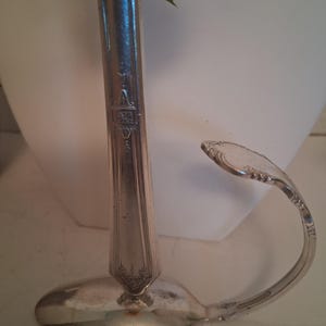 Könnte beinhalten: Eine silberfarbene Metall-Blumenvase aus wiederverwertetem Besteck. Die Basis besteht aus der Laffe eines Löffels, während der vertikale Stiel ein Griff mit eingravierten dekorativen Mustern ist. Ein gebogener Löffelstiel bildet einen seitlichen Henkel.