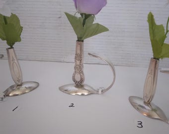 May include: Three silver-toned metal bud vases crafted from repurposed spoons, each featuring a slender handle stem and a wide bowl base. The center vase holds a single artificial lavender rose, while the outer vases hold white flowers.