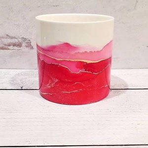 May include: A white ceramic cylinder vase with a red, pink, and gold abstract design. The vase has a smooth, glossy finish.