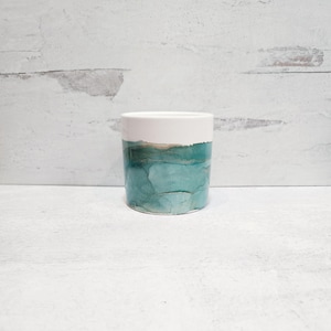 May include: A cylindrical ceramic container with a white upper section and a lower section featuring a teal and gold watercolor design. The container is set against a white background.