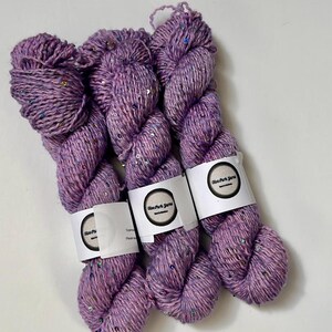 Peut inclure: Trois écheveaux de fil de couleur lavande avec des paillettes irisées. Chaque écheveau est enveloppé d'une étiquette blanche avec un logo noir. Le fil est disposé sur une surface blanche, mettant en valeur sa texture et ses variations de couleur.