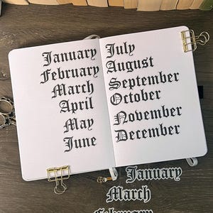 Könnte beinhalten: Ein aufgeschlagenes Notizbuch mit weißen Seiten, auf denen die Monate des Jahres von Januar bis Dezember in schwarzer gotischer Schrift gedruckt sind. Goldene Metall-Binderclips halten die Seiten fest. Drei weitere Monatsaufkleber liegen darunter.
