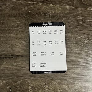 K&ouml;nnte beinhalten: Ein rechteckiger Aufkleberbogen mit der &Uuml;berschrift Tiny Tabs, der schwarz-wei&szlig;e Etiketten f&uuml;r Monate, notes, year, cal, daily und monthly enth&auml;lt. Der Bogen hat oben und unten einen schwarzen, gewellten Rand mit dem Text danicristine unten.