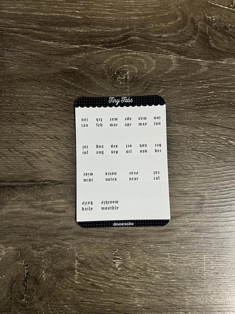 K&ouml;nnte beinhalten: Ein rechteckiger Aufkleberbogen mit der &Uuml;berschrift Tiny Tabs, der schwarz-wei&szlig;e Etiketten f&uuml;r Monate, notes, year, cal, daily und monthly enth&auml;lt. Der Bogen hat oben und unten einen schwarzen, gewellten Rand mit dem Text danicristine unten.