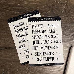 May include: Two white cards with black text and a scalloped border. The cards read "Cosmic Monthly" at the top, followed by the months of the year. The text is in a decorative font with star and plus symbols. The bottom right corner of the card has the text "danicacristine".