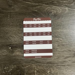 K&ouml;nnte beinhalten: Ein rechteckiger Bogen mit Aufklebern namens Tiny Tabs mit braunen und wei&szlig;en Reihen. Die Aufkleber enthalten Monate von Jan bis Dez, Beschriftungen f&uuml;r misc, notes, year, cal, daily und monthly sowie den Markennamen danicacristine am unteren Rand.
