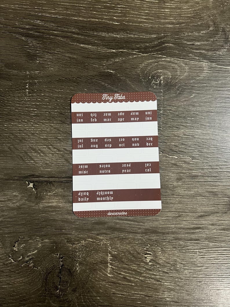 K&ouml;nnte beinhalten: Ein rechteckiger Bogen mit Aufklebern namens Tiny Tabs mit braunen und wei&szlig;en Reihen. Die Aufkleber enthalten Monate von Jan bis Dez, Beschriftungen f&uuml;r misc, notes, year, cal, daily und monthly sowie den Markennamen danicacristine am unteren Rand.