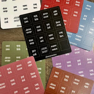 Könnte beinhalten: Eine Sammlung von Aufkleberbögen mit Monatsbezeichnungen, Jahresmarkierungen für 2027 und Notiz-Tabs in verschiedenen Farben wie Schwarz, Weiß, Rot, Blau, Grün und Braun. Jeder Bogen enthält vorgestanzte Tab-Formen mit aufgedrucktem Text.