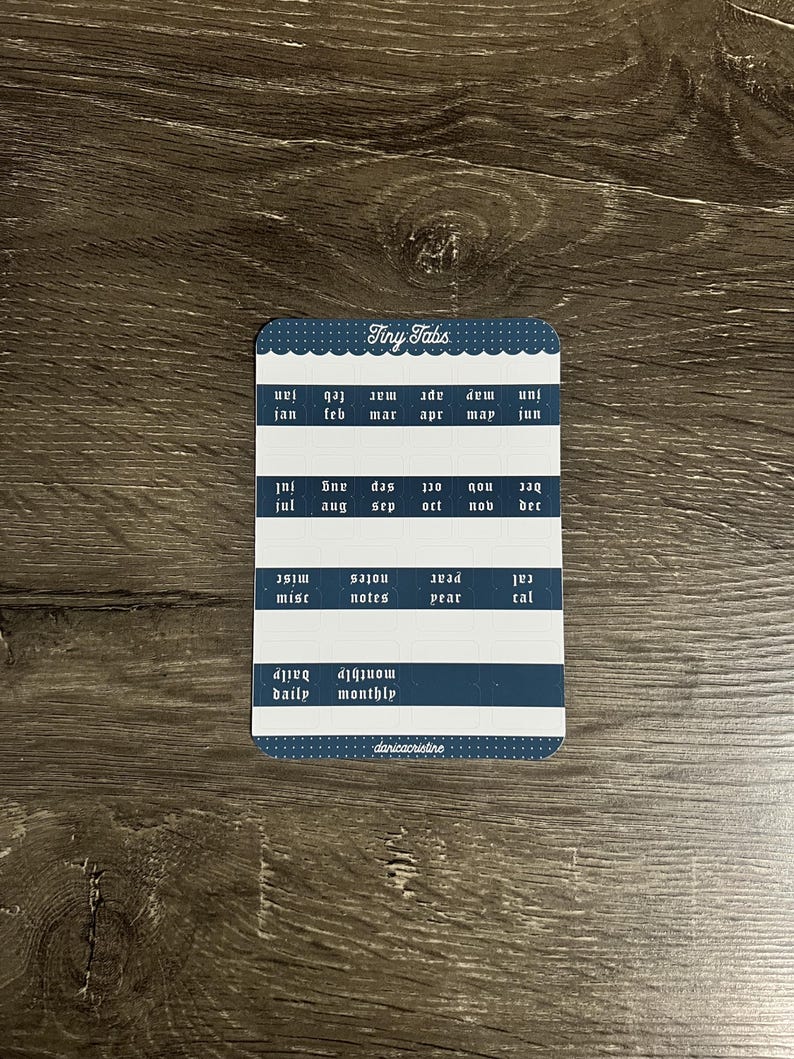 K&ouml;nnte beinhalten: Ein rechteckiger Aufkleberbogen mit der Aufschrift Tiny Tabs, dunkelblauem Hintergrund und wei&szlig;em Punktmuster am Rand. Er enth&auml;lt Reihen kleiner Klebelaschen, beschriftet mit den Monaten Jan bis Dez sowie misc, notes, year, cal, daily und monthly.