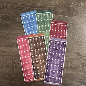 May include: A selection of planner stickers in various colours, arranged on a wooden surface. The rectangular stickers have rounded edges and feature a grid of dots and the days of the week. Colours include blue, green, red, brown, and purple.