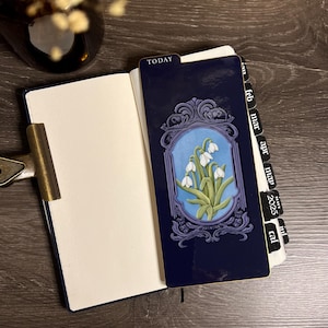 K&ouml;nnte beinhalten: Ein dunkelblaues Notizbuch mit einem dekorativen Lesezeichen mit floralem Design. Das Lesezeichen hat einen blauen Hintergrund mit wei&szlig;en Blumen und einem dekorativen Rahmen. Das Wort "TODAY" ist auf dem Lesezeichen zu sehen.