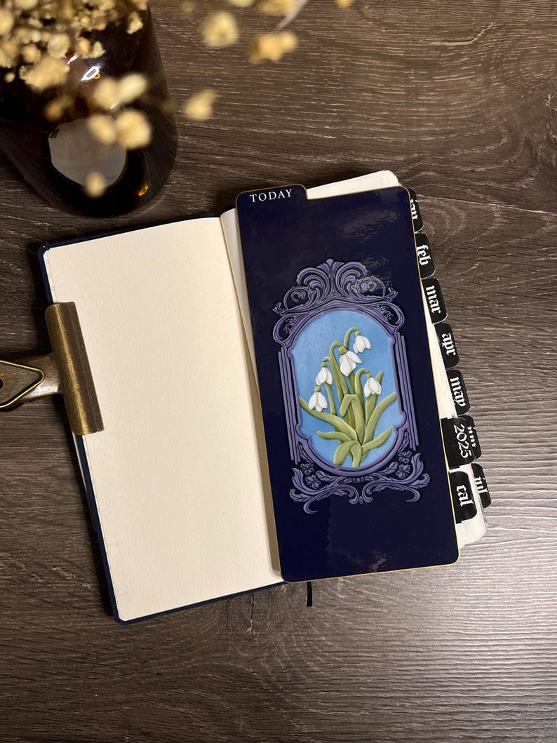 K&ouml;nnte beinhalten: Ein dunkelblaues Notizbuch mit einem dekorativen Lesezeichen mit floralem Design. Das Lesezeichen hat einen blauen Hintergrund mit wei&szlig;en Blumen und einem dekorativen Rahmen. Das Wort "TODAY" ist auf dem Lesezeichen zu sehen.