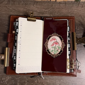 May include: Open leather planner with a burgundy cover and gold accents. The planner is open to a September calendar page and a decorative page with a floral design. The planner is on a wooden surface.