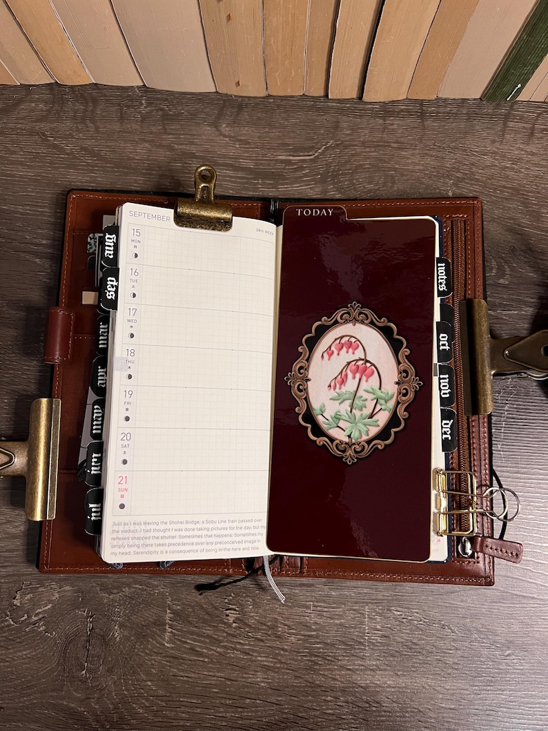 May include: Open leather planner with a burgundy cover and gold accents. The planner is open to a September calendar page and a decorative page with a floral design. The planner is on a wooden surface.