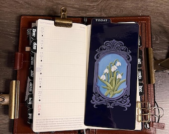 May include: A brown leather planner open to a September weekly spread with black month tabs. A dark blue bookmark features a white snowdrop flower illustration inside an ornate frame with the word TODAY printed at the top in white text.
