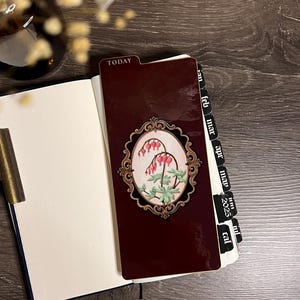 May include: A burgundy planner with a floral design and the word "TODAY" on the tab. The planner is open, revealing blank pages. A decorative bookmark with a floral design is inserted. The planner has monthly tabs for 2025.