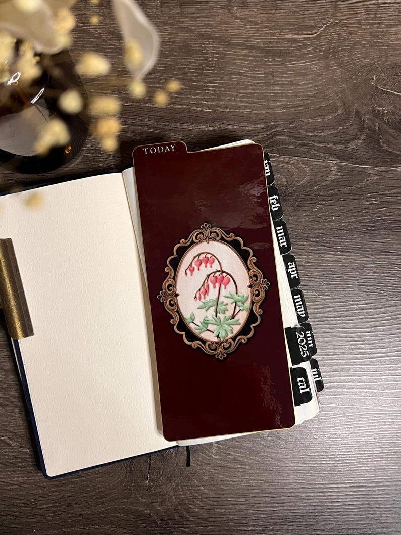 May include: A burgundy planner with a floral design and the word "TODAY" on the tab. The planner is open, revealing blank pages. A decorative bookmark with a floral design is inserted. The planner has monthly tabs for 2025.