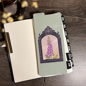 May include: Open notebook with a light blue cover and a gold-colored clip. The open pages reveal a light green divider with a purple floral illustration and the word "TODAY". Black tabs mark months and the year 2025.