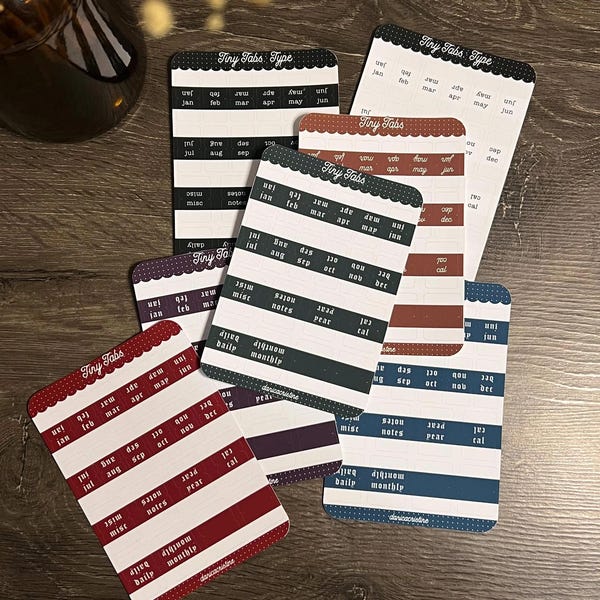 Puede incluir: Una colección de hojas de pegatinas rectangulares etiquetadas como Tiny Tabs y Tiny Tabs Type, que presentan filas de meses de enero a diciembre, junto con etiquetas para daily, monthly, notes y calendar. Las hojas vienen en negro, azul, rojo y morado con texto en blanco.