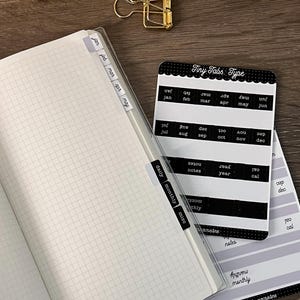 K&ouml;nnte beinhalten: Ein kariertes Notizbuch mit angebrachten Monats- und Kategorien-Tabs sowie ein Aufkleberbogen mit der Aufschrift Tiny Tabs Type, der schwarze und wei&szlig;e Etiketten f&uuml;r Monate, Notizen, Jahr und monatliche Eintr&auml;ge enth&auml;lt, plus zwei goldene Metallklammern.