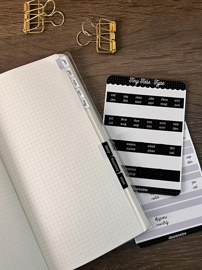 K&ouml;nnte beinhalten: Ein kariertes Notizbuch mit angebrachten Monats- und Kategorien-Tabs sowie ein Aufkleberbogen mit der Aufschrift Tiny Tabs Type, der schwarze und wei&szlig;e Etiketten f&uuml;r Monate, Notizen, Jahr und monatliche Eintr&auml;ge enth&auml;lt, plus zwei goldene Metallklammern.