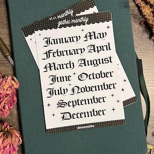 Könnte beinhalten: Ein weißer, rechteckiger Aufkleberbogen mit dem Text "gothic monthly" am oberen Rand und einer Liste aller zwölf Monate des Jahres in einer schwarzen gotischen Schriftart. Der Bogen ist mit kleinen schwarzen Sternsymbolen und einem gepunkteten Rand verziert.