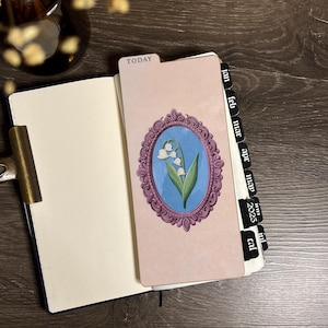 Puede incluir: Un marcapáginas para agenda con fondo rosa claro que presenta una ilustración central de lirios de los valles blancos dentro de un marco ovalado morado ornamentado. El marcapáginas tiene una pestaña etiquetada TODAY y pestañas negras para 2025.