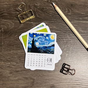 Puede incluir: Una pila de pequeños calendarios cuadrados con la pintura "Noche estrellada" de Van Gogh. El calendario muestra el mes de enero con la fecha 01. Un clip dorado, un clip negro y un lápiz están sobre una superficie de madera.