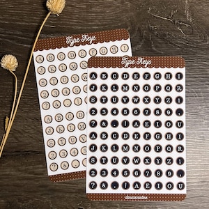 Könnte beinhalten: Zwei rechteckige Aufkleberbögen mit der Aufschrift Type Keys, die Reihen von kreisförmigen Aufklebern mit Buchstaben, Zahlen und Satzzeichen enthalten. Die Aufkleber haben einen dunkelbraunen Hintergrund mit weißem Text und weißem Rand. Die Bögen haben braune Ränder mit weißen Punkten.