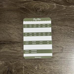 K&ouml;nnte beinhalten: Ein rechteckiger Aufkleberbogen mit abgerundeten Ecken und der Aufschrift Tiny Tabs, der olivgr&uuml;ne Zeilen und wei&szlig;en Text aufweist. Die Etiketten umfassen die Monate Jan bis Dez sowie misc, notes, year, cal, daily und monthly. Der Text ist in zwei Ausrichtungen gedruckt.