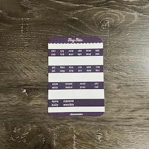 K&ouml;nnte beinhalten: Ein rechteckiger Bogen mit Klebeetiketten namens Tiny Tabs in einem violett-wei&szlig;en Farbschema. Der Bogen enth&auml;lt Reihen mit Monatsnamen, Kategorien wie misc, notes, year, cal, daily, monthly sowie den Schriftzug danicacristine.