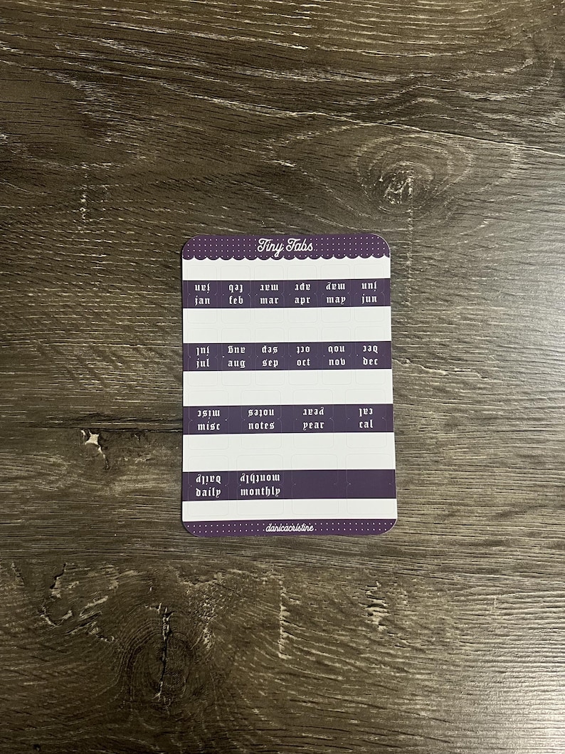 K&ouml;nnte beinhalten: Ein rechteckiger Bogen mit Klebeetiketten namens Tiny Tabs in einem violett-wei&szlig;en Farbschema. Der Bogen enth&auml;lt Reihen mit Monatsnamen, Kategorien wie misc, notes, year, cal, daily, monthly sowie den Schriftzug danicacristine.