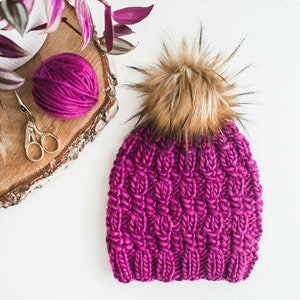 Pode incluir: Um gorro de malha rosa com um pompom de pele sintética marrom. O gorro é feito com um fio grosso e tem um padrão de malha trançada.