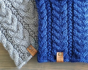 May include: Two cable-knit beanies in grey and blue. The beanies have a textured, woven appearance with a leather tag attached. The tags have the text "Urban Chic Crafts".