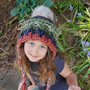 May include: A child wearing a green, blue, and red knitted hat with a white pom pom and matching long tassels. The hat has a chunky knit texture. The child is wearing a dark grey t-shirt with gold lettering that says "always be a good mom". The child is sitting on a green surface.