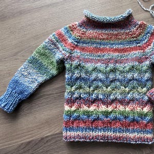 May include: A colorful knitted sweater with a high neck and long sleeves. The sweater has a striped pattern in shades of blue, green, red, and white.