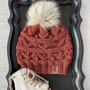 Op de afbeelding: Een roestkleurige gebreide beanie met een pluche witte pompon. De beanie heeft een kabelbreipatroon en een bruin leren label met een hert-silhouet.