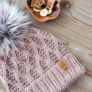 Op de afbeelding: Mauve gebreide beanie met een imitatiebont pompon. De hoed heeft een textuurpatroon en een klein leren label met de tekst "Urban Chic Crochet". Houten knopen en labels liggen in een houten kom.