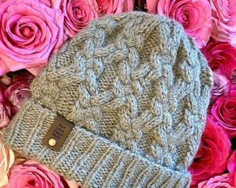 May include: A gray knitted beanie with a brown leather tag that reads "Knit Love Hand Knits". The beanie is surrounded by pink roses.